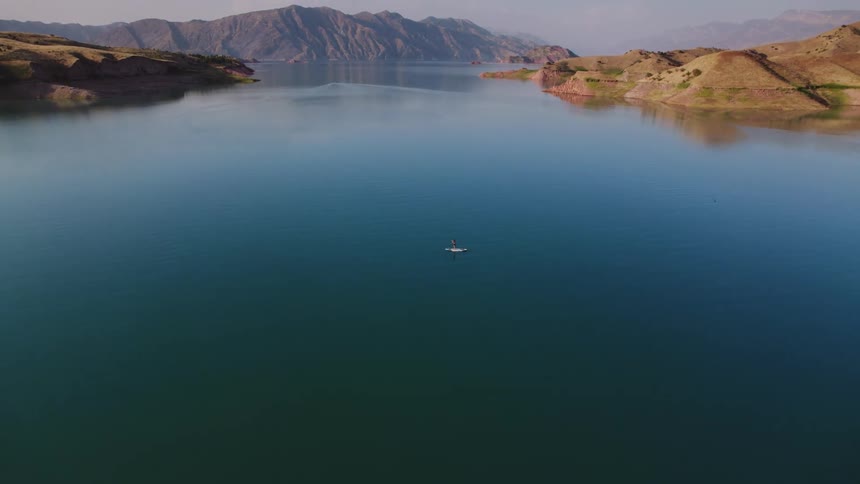 An aerial shot of a calm body of water surrounded by mountains offers an initially wide vista that moves downwards centering a person on a paddleboard