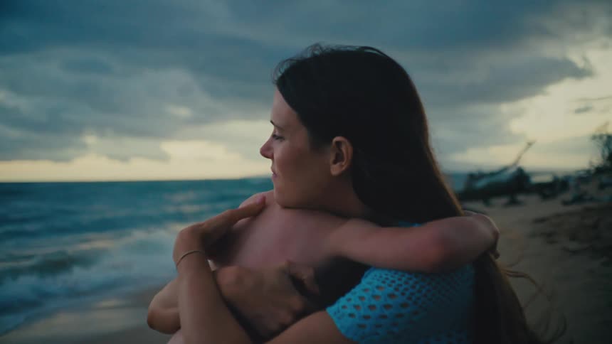 Mother and son embrace each other with waves lapping the beach behind them