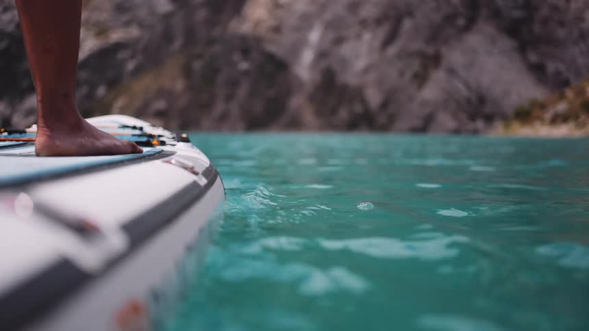 The person is paddling while standing on a surf board going towards a cliff in front of them