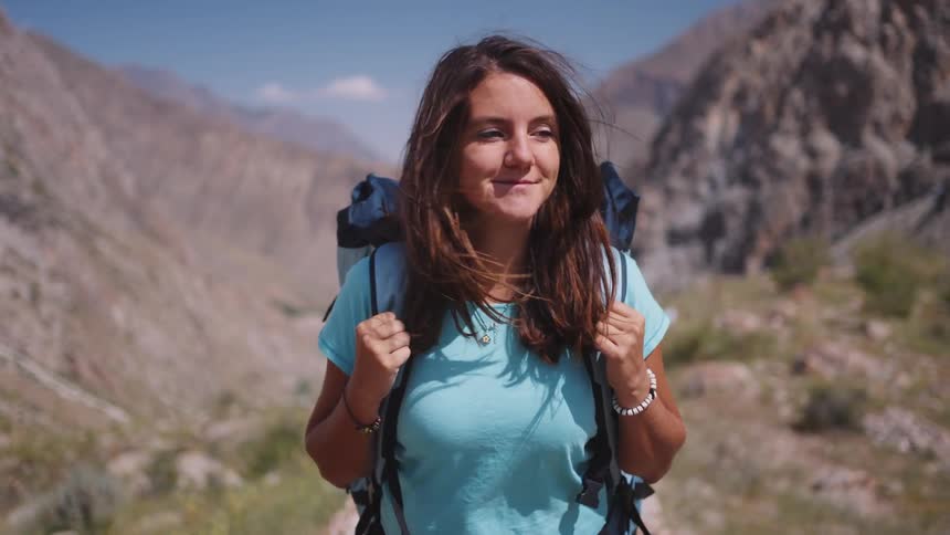 A young woman with long brown hair wearing a blue shirt and a backpack walks against a backdrop of rocky mountains appearing to be on a hiking adventure