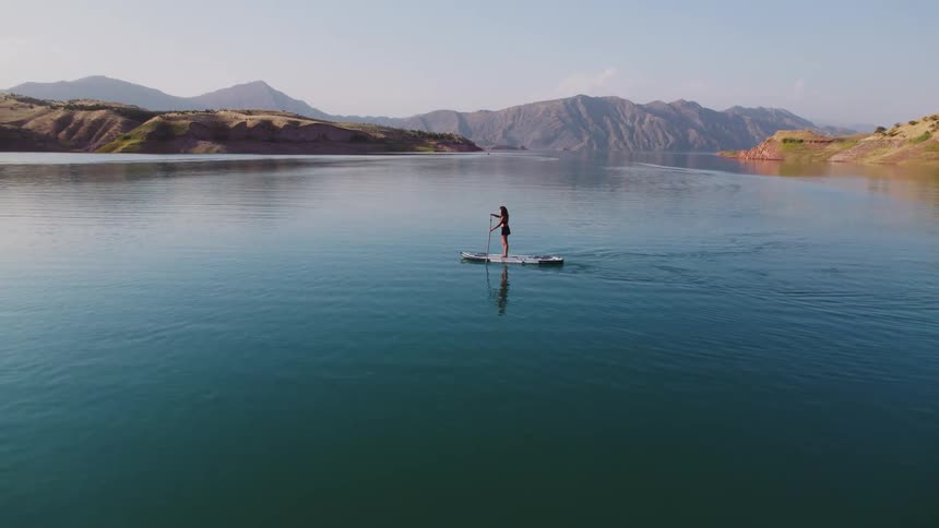 A lone figure stand up paddleboards on a calm expansive lake surrounded by rolling hills and mountains under a clear sky