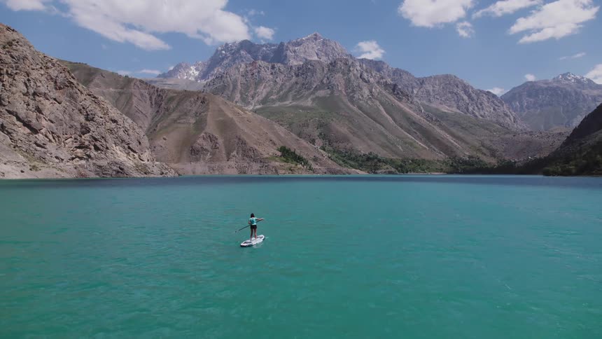 An aerial view shows a woman standup paddleboarding across a vibrant blue mountain lake surrounded by towering peaks