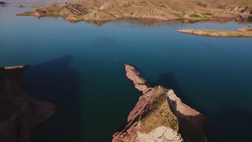 A highangle aerial view displays a serene lake surrounded by arid hills with a rocky headland extending into the calm water