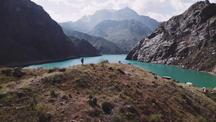 A lone figure stands on a hilltop overlooking a turquoise lake nestled between towering mountains