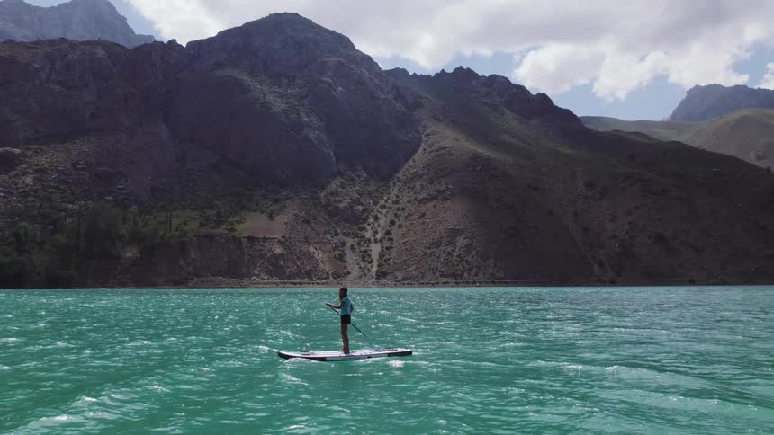 A woman stands on a paddleboard in vibrant turquoise water surrounded by rugged mountains under partly cloudy skies as gentle waves ripple around her while she propels herself forward maintaining balance