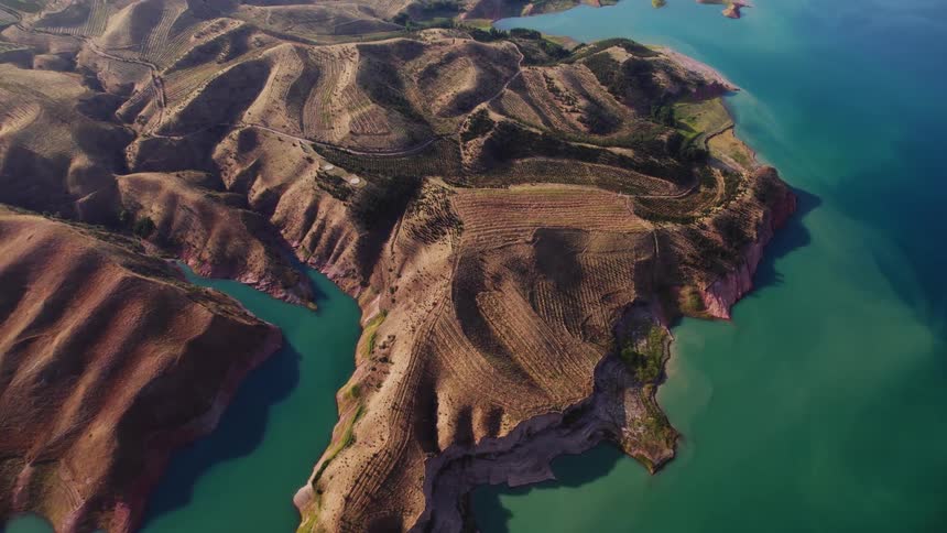 A sweeping aerial perspective showcases a rugged coastline meeting turquoise water characterized by rolling hills and complex geographical features