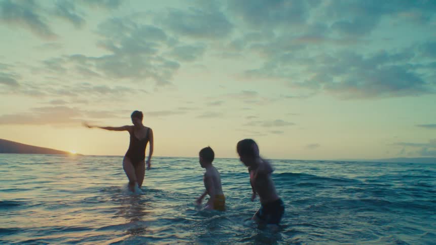 Mother playing with two young sons in shallow ocean surf at tropical beach during sunset