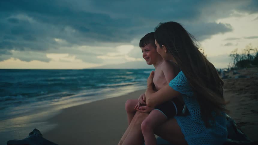 Mother kissing laughing young son on the cheek while sitting on tropical beach at sunset