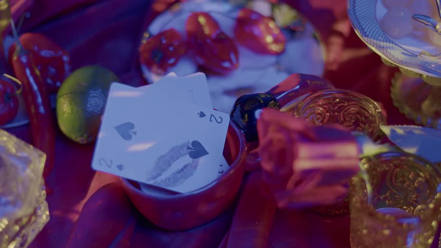 A static closeup exhibits a tabletop arrangement with various objects like fruit playing cards and glassware with dynamic lighting is projecte on to it
