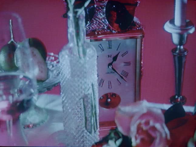 A closeup of a still life arrangement featuring a clock glassware and fruit is presented as the brightness changes and the light is project on to it