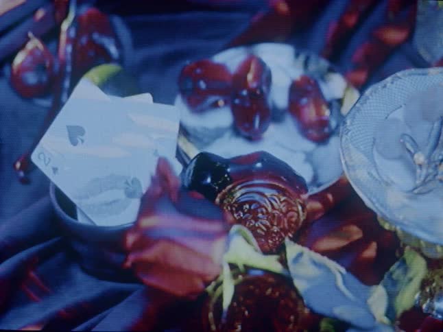 A close up reveals a two of spades playing card nestled in a bowl surrounded by small glass objects illuminated by dynamic shifting red lights