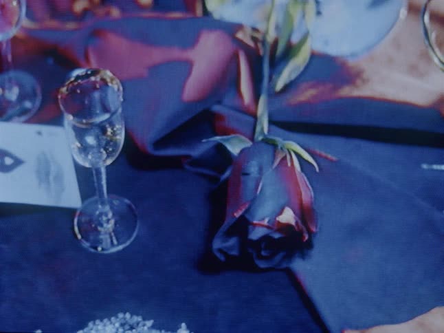 A static closeup features a single red rose and a wine glass on a tabletop dramatically illuminated by projected light within surrounding darkness