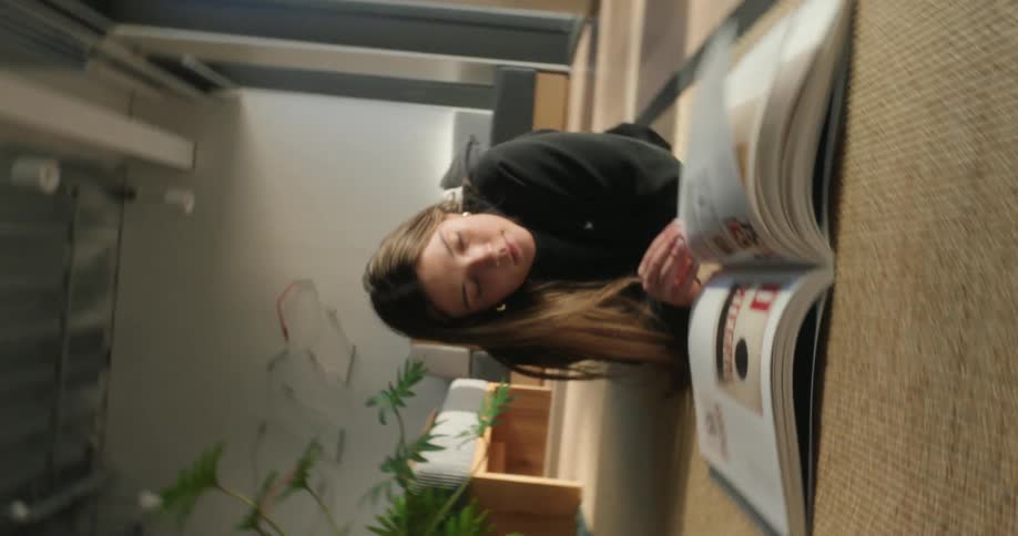 A woman lies on the floor reading a book in a minimalist room near large windows