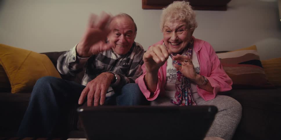 An older couple sits together on a couch in a living room cheerfully waving at a tablet while engaged in a video call
