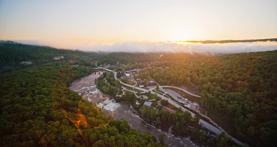 Orbiting over a small river town surrounded by dense forest at sunset revealing roads bridges and a waterfall with golden light spilling across the landscape and low clouds hugging the distant hills