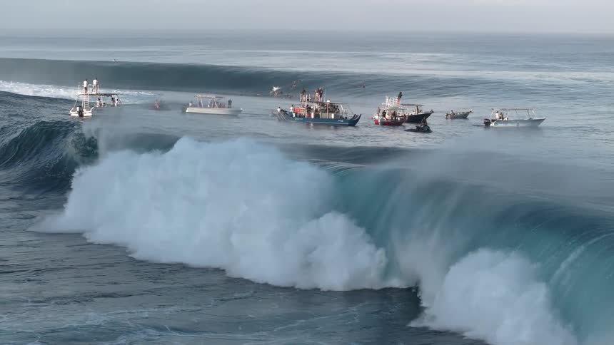 People fill several boats and watch surfers while waves swell and break on a slightly foggy day