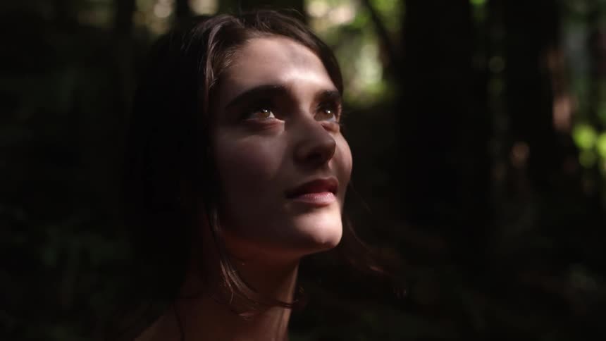 Woman stands in a forest looking around as rays of sun hit her face
