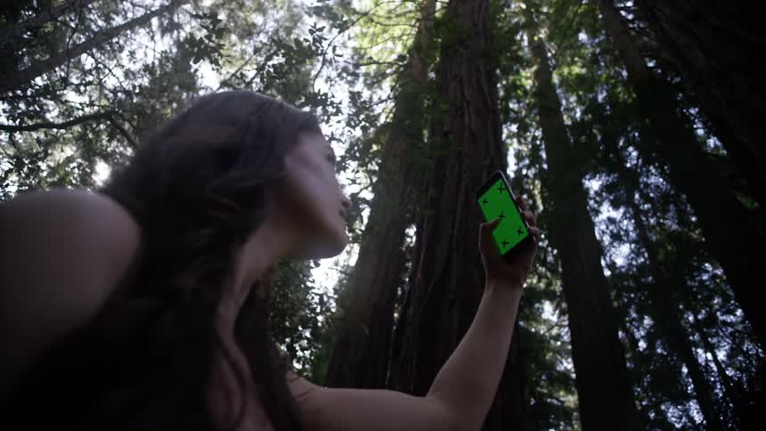 A woman holding a phone and spinning around in the woods looking for a signal