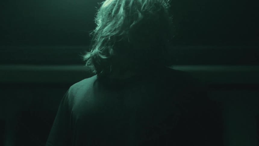 A long haired drummer in a dimly lit room puts on a black t shirt and walks away from the drum set