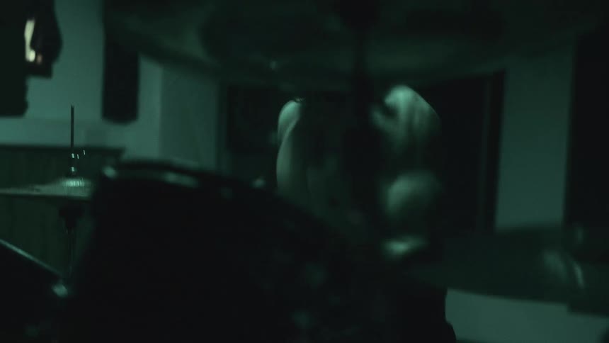 A muscly man stops to take his shirt off as he plays the drums