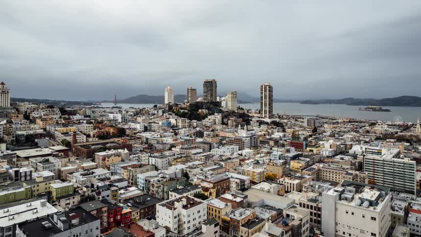 A densely packed cityscape with a mix of low rise buildings and taller skyscrapers stretching towards a bay with islands and a bridge in the distance under an overcast sky