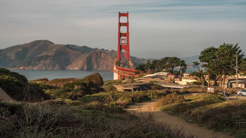 The golden gate bridges tower rising above a landscape of coastal vegetation and buildings with a bay and hills in the background