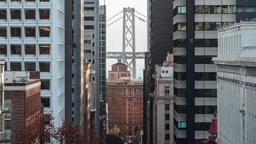A timelapse captures cars driving across the san franciscooakland bay bridge and down a city street below with tall buildings framing the view and flags rapidly waving between them