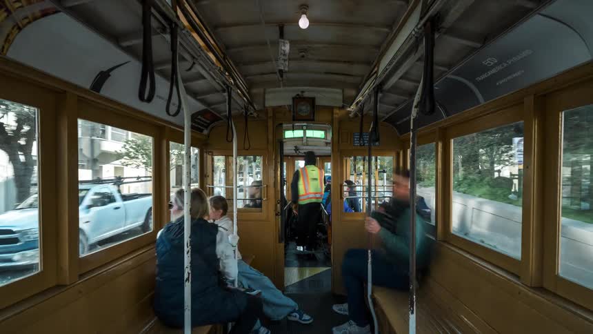 The interior of a vintage streetcar is shown with passengers seated on wooden benches and a conductor in a reflective vest standing near the doorway as the vehicle moves