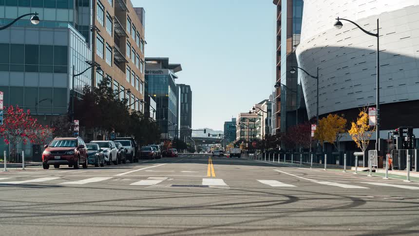 A wide urban street lined with modern buildings and trees featuring moderate traffic and clear crosswalk markings under a bright sky