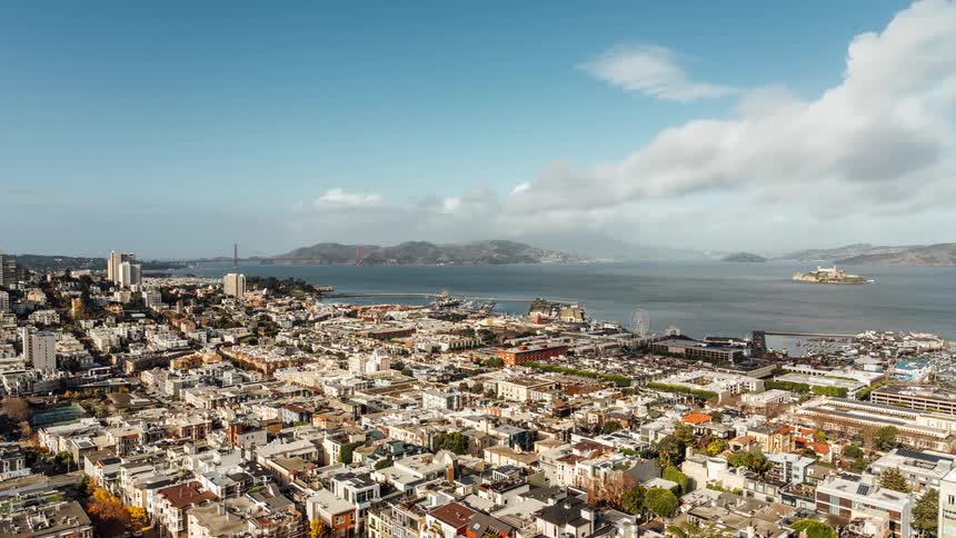 Aerial view of a sprawling cityscape with densely packed buildings a waterfront area and a body of water in the distance all under a blue sky with scattered clouds