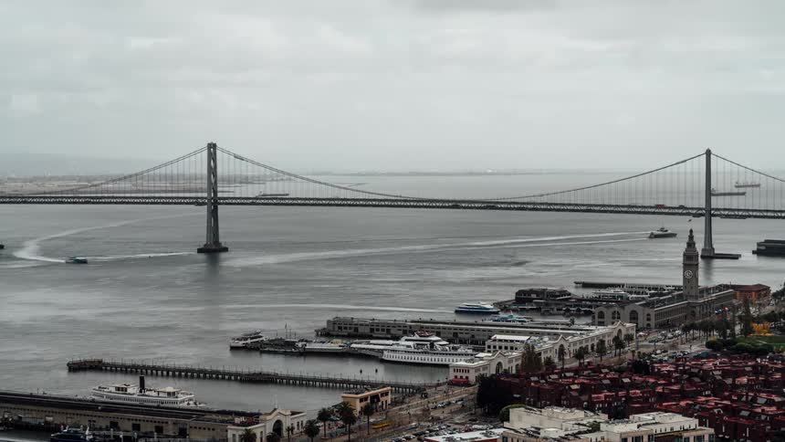 An aerial timelapse with tiltshift effect shows ferries and boats moving quickly around the san francisco ferry docks with the bay bridge spanning the water on an overcast day