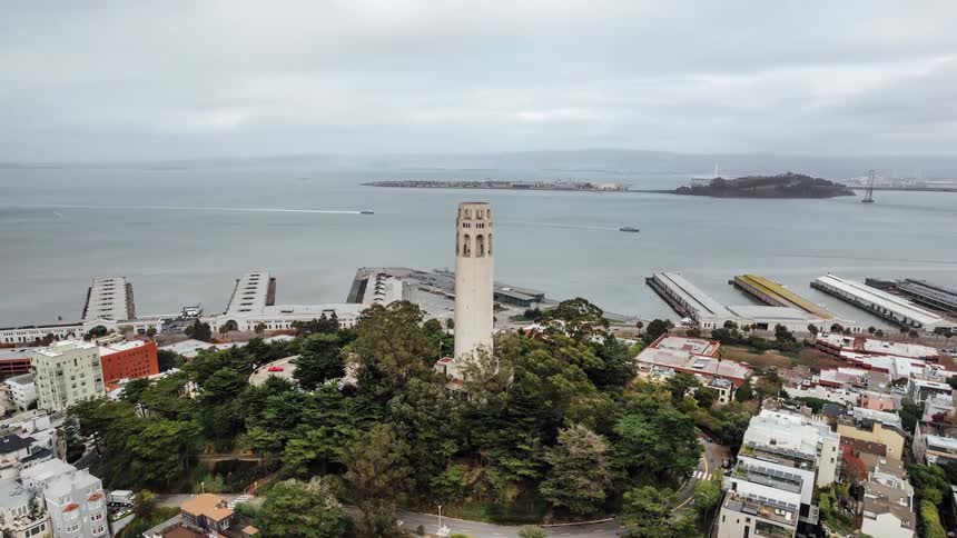 An aerial view shows coit tower standing prominently on a hill overlooking san franciscos cityscape with a bay and a bridge visible in the background