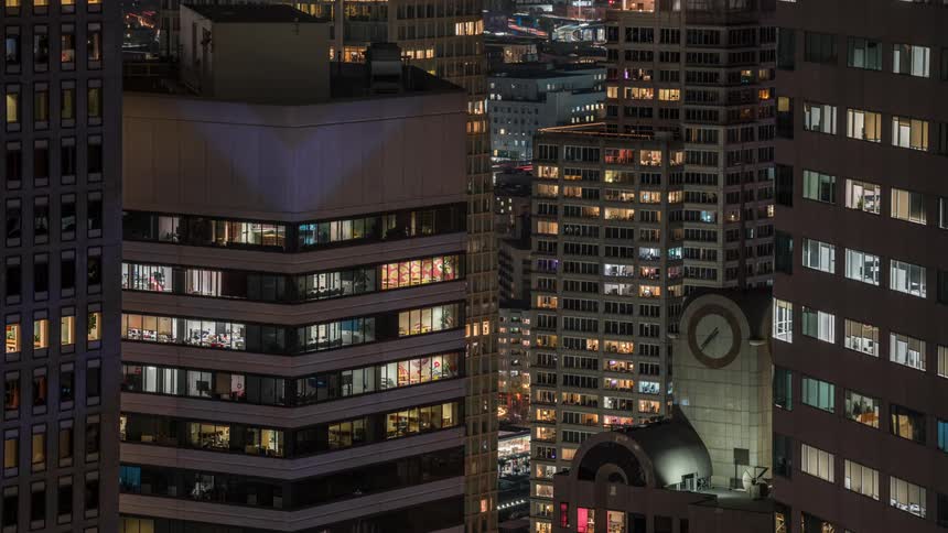 An aerial view shows multiple tall buildings at night with illuminated windows creating a grid like pattern and a clock face visible on one of the buildings