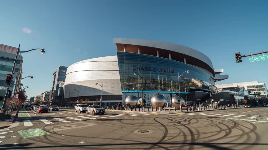 The chase center a modern arena with a curved glass facade stands beside a city street with crosswalks and passing vehicles under a clear sky