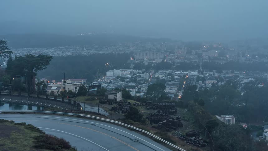 A winding road in the foreground leading to a sprawling city with illuminated buildings and a distant bridge spanning across water against a backdrop of hills