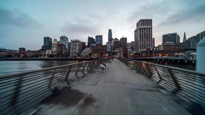 A wet concrete pier stretches toward the illuminated san francisco skyline at dusk flanked by calm water and docked boats