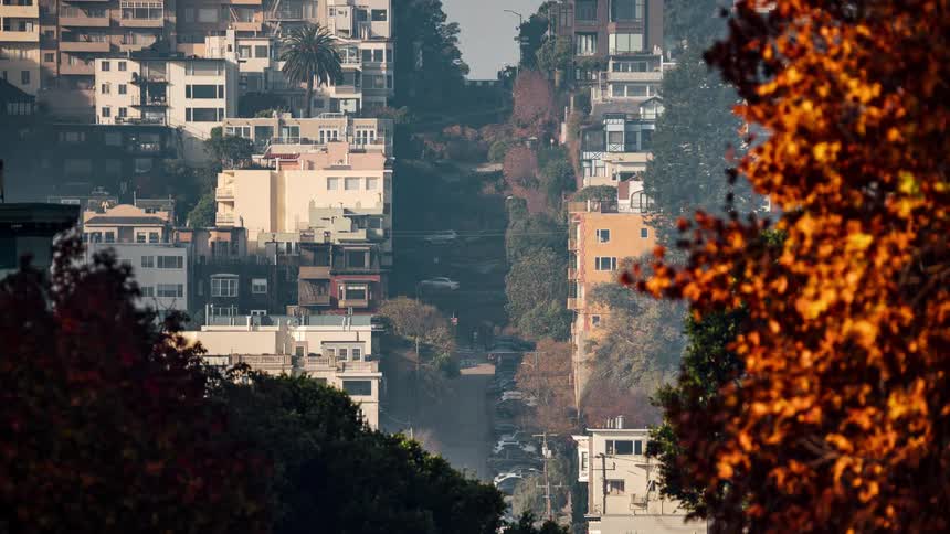 A timelapse captures cars navigating the sharp curves of san franciscos lombard street viewed from a distance and framed by trees with autumn foliage