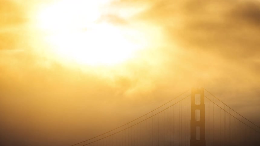 The sun bursts brilliantly through dense fog enveloping the top of a golden gate bridge tower in this closeup timelapse shot bathing the scene in a warm hazy light