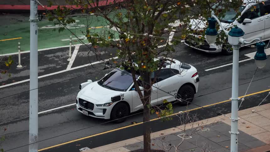 A selfdriving white vehicle with a prominent sensor rig on its roof navigates an urban street with painted bike lanes and tram tracks