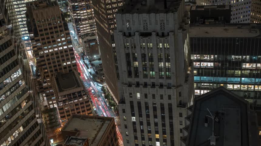 An aerial view shows a tall ornate building among other city structures at night with streetlights and car trails visible below