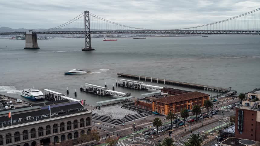 An aerial view shows the bay bridge spanning across the water with piers and buildings along the waterfront under an overcast sky