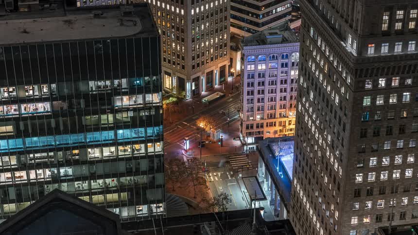 A city intersection at night framed by illuminated skyscrapers and streetlights with blurred car light trails indicating movement