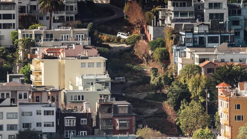 A densely packed neighborhood is shown with a mix of architectural styles situated on a hillside with cars driving down a winding street