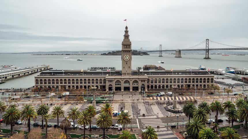 An aerial view shows the san francisco ferry building with its clock tower surrounded by palm trees and a plaza with the bay bridge in the background