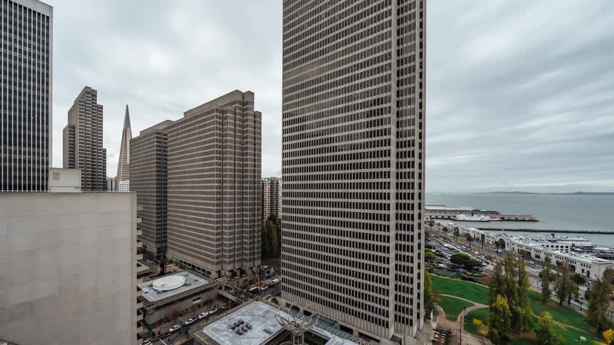 An elevated view captures a cluster of tall buildings in an urban setting with a glimpse of the bay and piers in the distance under an overcast sky