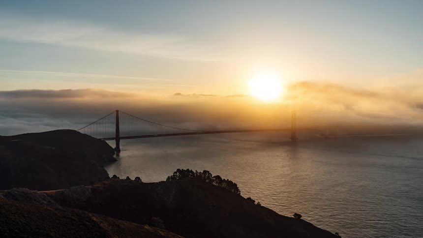 An aerial view shows the golden gate bridge stretching across the frame partially obscured by fog with the sun setting in the distance