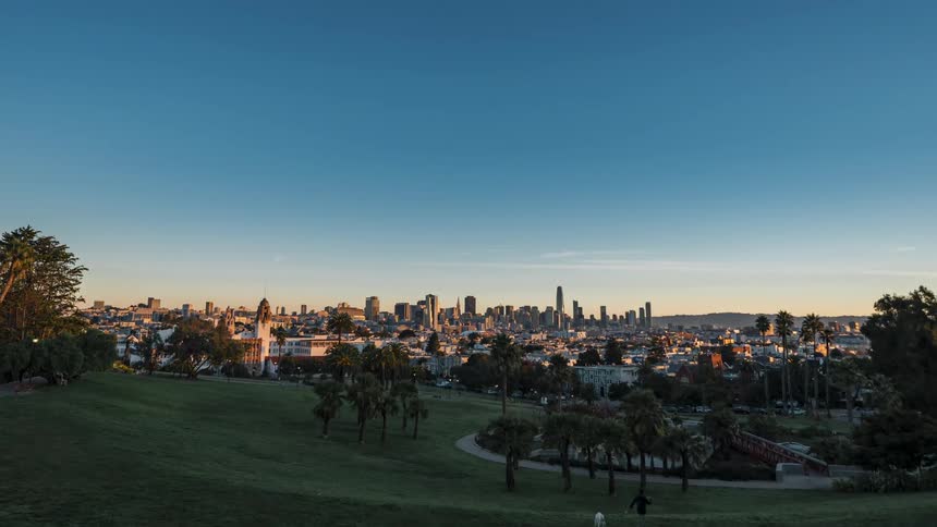 A cityscape taken from a slightly elevated position featuring a park with trees and streetlights in the foreground and a distant skyline illuminated against a dark blue sky