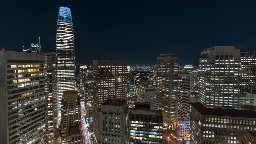 A nighttime aerial view captures the illuminated salesforce tower a prominent skyscraper with a distinctive segmented pattern of lights towering over a dense urban landscape of other lit buildings and city streets against a dark clear sky