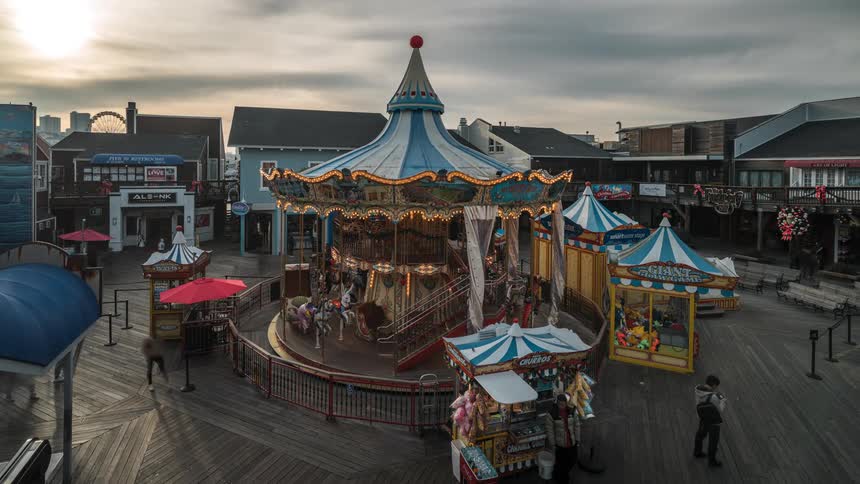 A carousel with blue and white striped canopies stands on a wooden pier surrounded by shops under an overcast sky