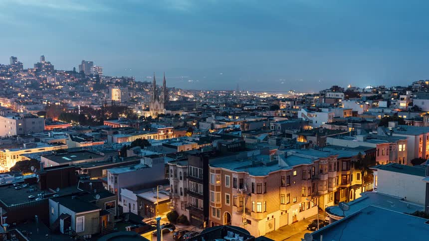 A panoramic view of a dense urban cityscape with closely packed buildings featuring a church steeple rising above the rooftops and a hazy skyline in the distance